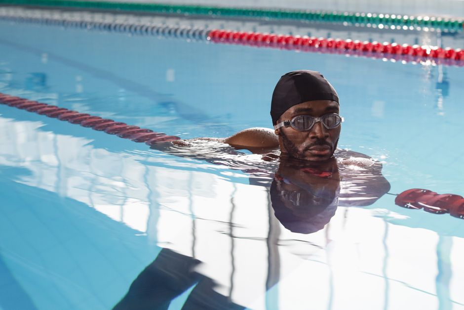 A male swimmer with dark skin, wearing a black swim cap and clear goggles, is positioned in a swimming pool, resting his arms on the edge of the pool. The pool has clear, blue water with gentle ripples, and the surface reflects the surrounding structure, including white light and metal framework. The swimmer is looking straight ahead with a focused expression. In the background, there are red and blue lane dividers stretching across the pool, and the environment suggests an indoor aquatic facility with natural light filtering through large windows, creating soft reflections on the water's surface. The scene depicts a moment of rest during a swimming session or training, with the pool area maintained and clean, consistent with typical facility standards.