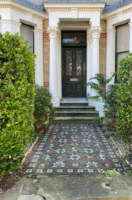 A close-up view of a traditional front garden entrance featuring a patterned stone pathway leading up to a black front door framed by white columns and decorative molding. The pathway is composed of small, geometric tiles in shades of black, white, and grey, creating a mosaic-like design. On either side of the pathway, lush, well-maintained green shrubs and bushes border the garden, with the greenery contrasting against the light-colored exterior wall of the house. The steps leading to the door are made of grey stone, and the porch area is slightly elevated. The environment appears clean and tidy, with natural daylight illuminating the scene, highlighting the textures of the plants, pathway, and architectural details. This scene exemplifies careful garden maintenance and landscaping work by Gardeners Dulwich, focusing on curb appeal and front garden aesthetics suitable for a residential property in Lordship Lane.